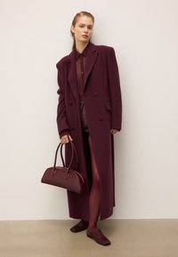 Femme portant un long manteau bordeaux, un haut assorti, des collants et des chaussures, tenant un sac à main bordeaux, se tenant contre un mur clair.