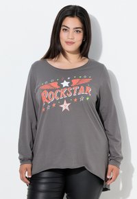 Kobieta z długimi ciemnymi włosami, ubrana w szary, długi rękaw z napisem "ROCKSTAR" oraz grafikami gwiazd i skrzydeł, w połączeniu z czarnymi spodniami.