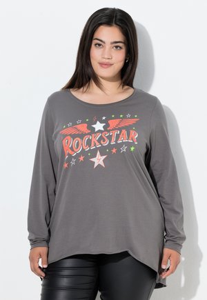 Vrouw met lang, donker haar die een grijze longsleeve-shirt draagt met de tekst "ROCKSTAR" en sterren- en vleugelgraphics, gecombineerd met zwarte broek.