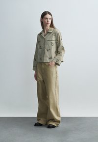 Femeie stând cu mâna în buzunar, purtând o jachetă bej cu dungi florale și pantaloni largi bej cu mici modele punctate pe un fundal simplu.