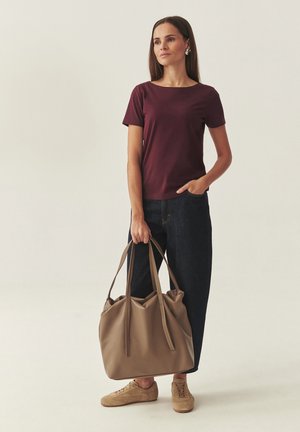 Kvinde iført en bourgogne rød kortærmet skjorte, mørke jeans og beige sneakers, der holder en stor lysebrun skuldertaske mod en ensfarvet baggrund.