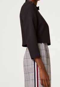 Veste noire courte avec un col rond, ornée d'un petit nœud. Portée sur une jupe à carreaux avec des rayures verticales rouges et blanches.