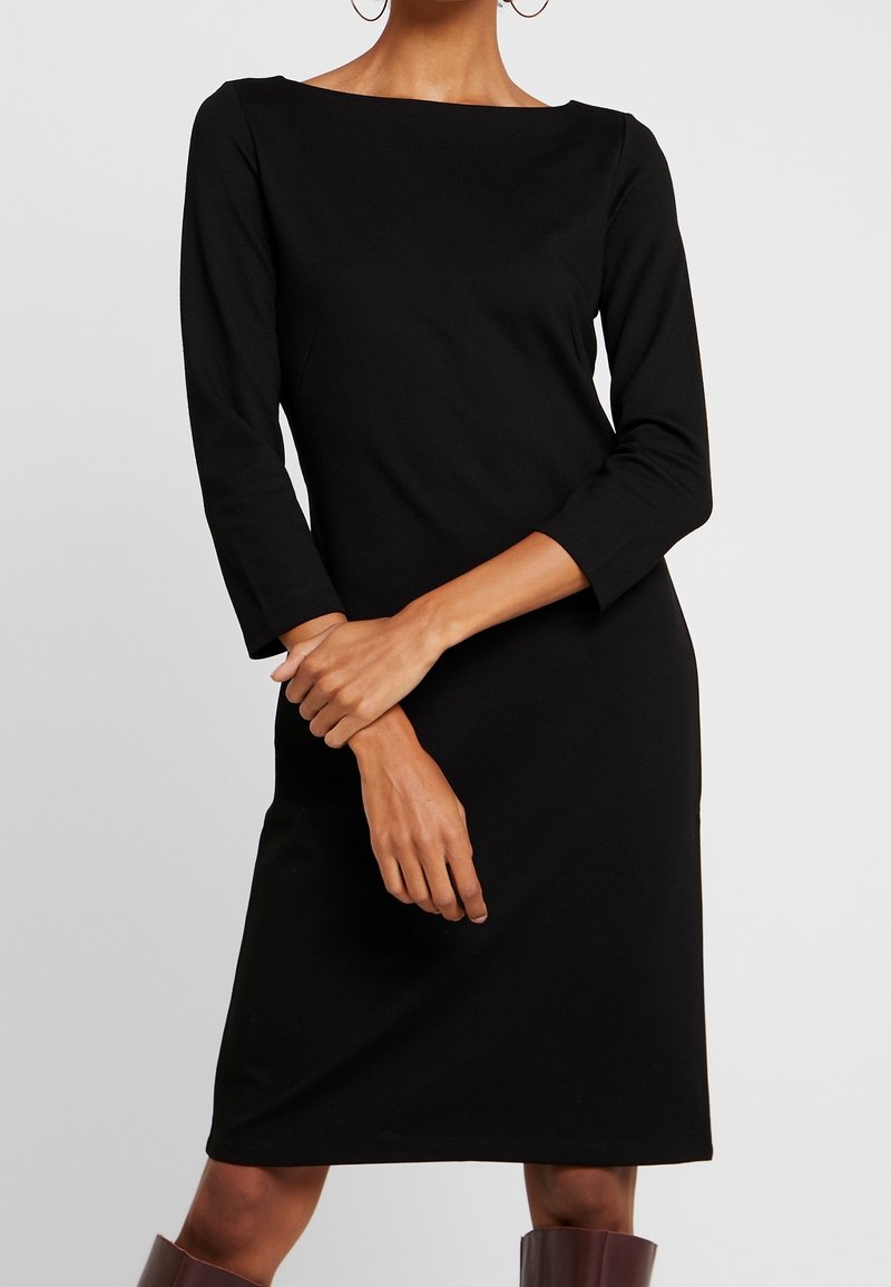Femme portant une robe noire simple, longueur genou, manches trois-quarts, et des bottes marron montant jusqu'aux genoux sur un fond uni.
