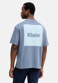 Camiseta de manga corta gris jaspeado con un gran cuadrado azul claro y el texto "Klein" en la parte trasera. Material de algodón suave, ajuste relajado.