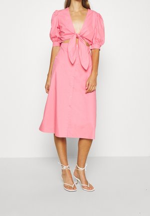 Femme portant une robe midi rose avec manches bouffantes, détail nœud à l'avant, découpes à la taille, et sandales blanches à talons hauts à brides.