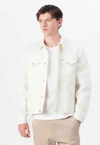 Hvid denimjakke med to brystlommer, sølvknaplukninger og stående krave, båret over en hvid T-shirt og beige bukser.