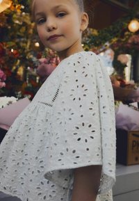 Robe blanche en broderie anglaise avec découpes florales, manches courtes et texture froncée, sur un fond de fleurs colorées et de feuillage.