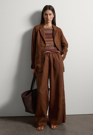 Junge Frau trägt braune Hosen mit weitem Bein, passendem Blazer, mehrfarbigem Strickpullover, braunen Sandalen und hält eine große, gewebte braune Tragetasche.