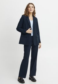 Tumši zilas krāsas uzvalks, kas sastāv no pielāgota žaketes ar iecirstiem uzvalka atlokiem, divām priekšējām kabatām un atbilstošām platām biksēm. Tekstūrēta, gluda auduma.