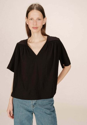 Blouse noire à col en V avec des manches courtes, en tissu léger, ornée de doux plis le long des épaules. Portée avec un jean bleu clair.