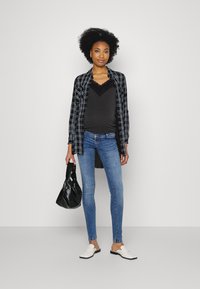 Schwarz-weißer kariertem Hemd, schwarzes Spitzen-Tanktop, blaue Skinny-Jeans, weiße Slipper, hält eine schwarze Tasche. Glatte Texturen, lässiger Schnitt.