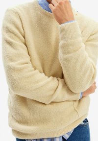 Pull en polaire couleur crème avec col rond et manches longues, doté d'une texture douce et de poignets côtelés, superposé à une chemise rayée bleue.