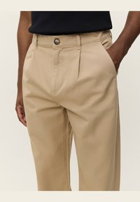 Tan, pleated trousers with a smooth texture. Features a button closure and side pockets, tailored fit, suitable for casual or smart attire.