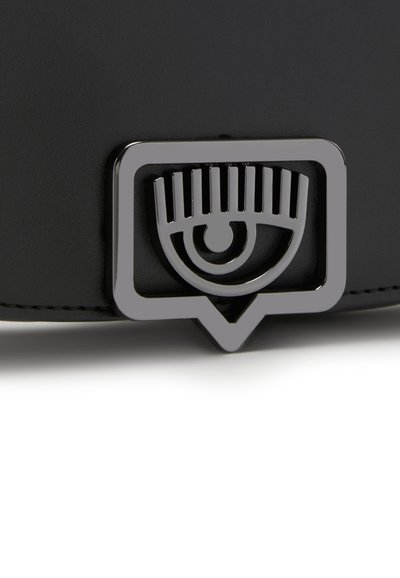 Accessoire en cuir noir avec un emblème métallique argenté en forme d'œil, orné de lignes verticales, encadré dans un rectangle.