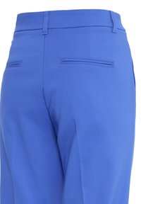 Vue de dos d’un pantalon bleu sur mesure avec passants de ceinture et deux poches arrière horizontales zippées.