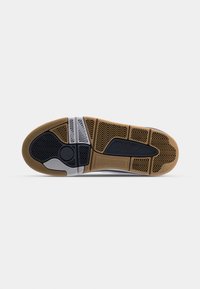 Semelle d'une basket avec des parties en caoutchouc marron, noir et gris, comportant différents motifs de crampons pour l'adhérence et la flexibilité.