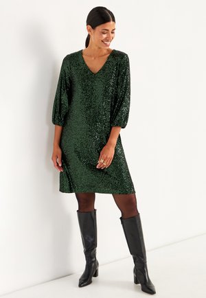 Grön paljetklänning med V-hals, puffärmar, knälång längd, ihop med svarta knähöga klackstövlar och texturerade svarta strumpbyxor.