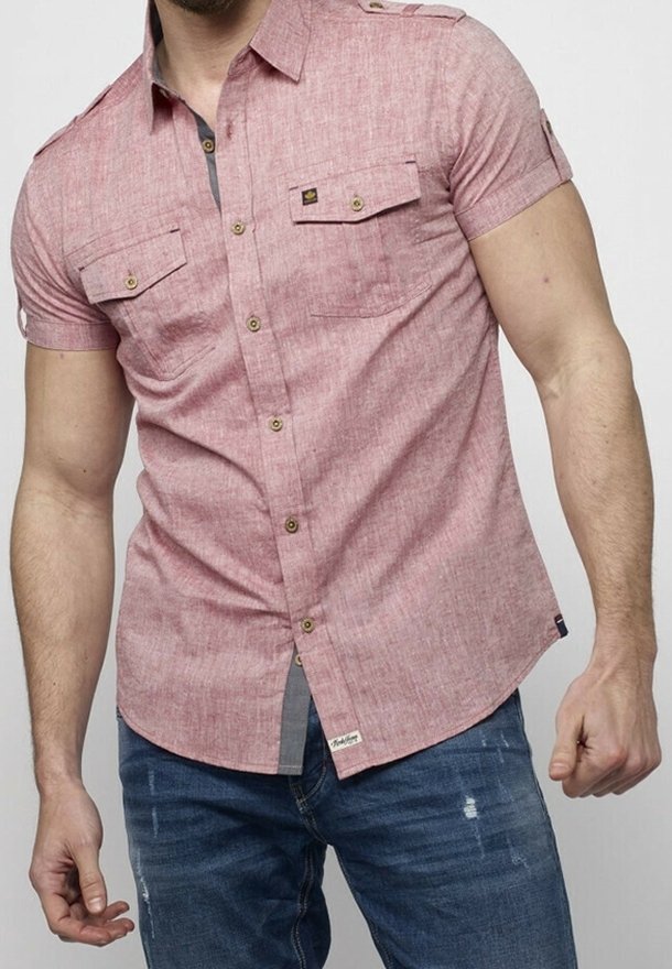 Homme portant une chemise rose à manches courtes boutonnée avec deux poches poitrine et un jean bleu délavé sur un fond uni.