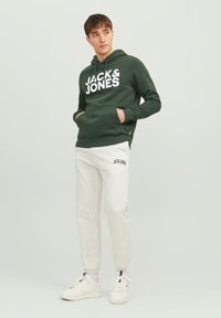 Green hoodie with white "JACK & JONES" logo, paired with light gray sweatpants with matching logo, and white sneakers. Casual, relaxed fit.
