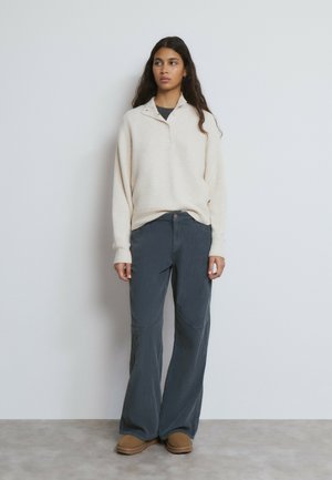 Pull à col en maille côtelée crème avec un bouton, associé à un pantalon évasé en velours côtelé gris foncé et des chaussons isolants beige.