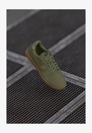 Sapatilha Nike verde com sola de borracha e swoosh texturizada, exibida sobre uma superfície de grelha metálica.