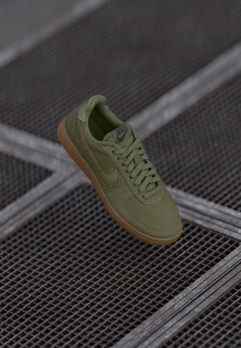 Zaļš Nike sneakers ar gumijas zoli un struktūrētu swoosh, izstādīts virs metāla režģa virsmas.