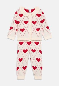 Krémově zbarvený pletený dvoudílný set obsahující kardigan se volánkovým límcem, červenými vzory srdcí a odpovídající kalhoty s manžetami.