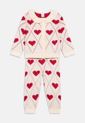 Ensemble deux pièces tricoté couleur crème comprenant un cardigan à col volanté, motifs de cœurs rouges, et un pantalon assorti avec des poignets.
