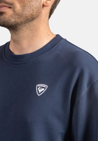 Felpa blu navy, in misto cotone, con collo a coste, caratterizzata da un distintivo bianco sul lato sinistro del petto. Tessuto morbido, vestibilità comoda.