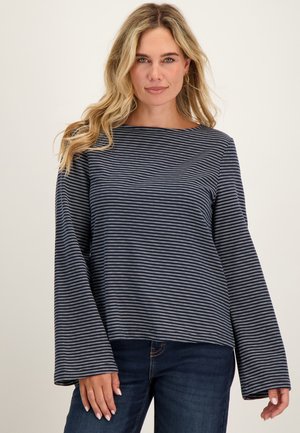 Vrouw met los blond haar die een marineblauw-wit gestreept shirt met lange mouwen en donkerblauwe spijkerbroek draagt, staand tegen een effen achtergrond.