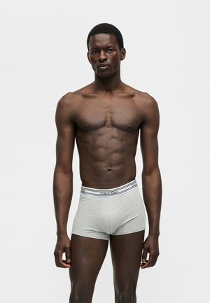 Άνδρας μοντέλο στέκεται μπροστά σε λευκό φόντο φορώντας ανοιχτό γκρι μποξεράκια Calvin Klein με επώνυμη μέση.