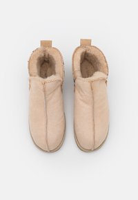Pantofole di lana beige con una morbida fodera in pile, caratterizzate da un design con cuciture a punti e una piccola linguetta sul retro per facilitare l'indossamento.