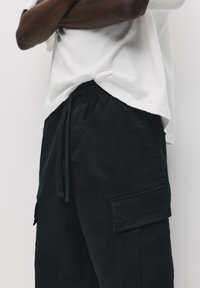 Zwarte cargobroek met een tailleband met trekkoorden, zijzakken en zichtbare stikdetails, gecombineerd met een wit shirt met korte mouwen.