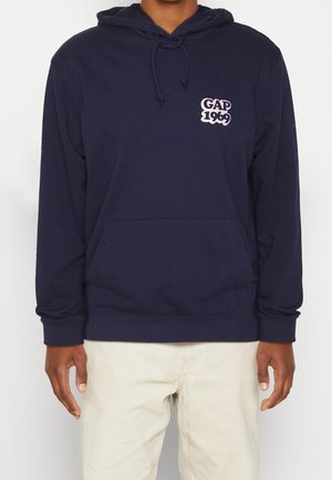 Person iført en mørkeblå hættetrøje med frontlomme og "GAP 1969" logo på brystet, kombineret med lysebeige bukser.