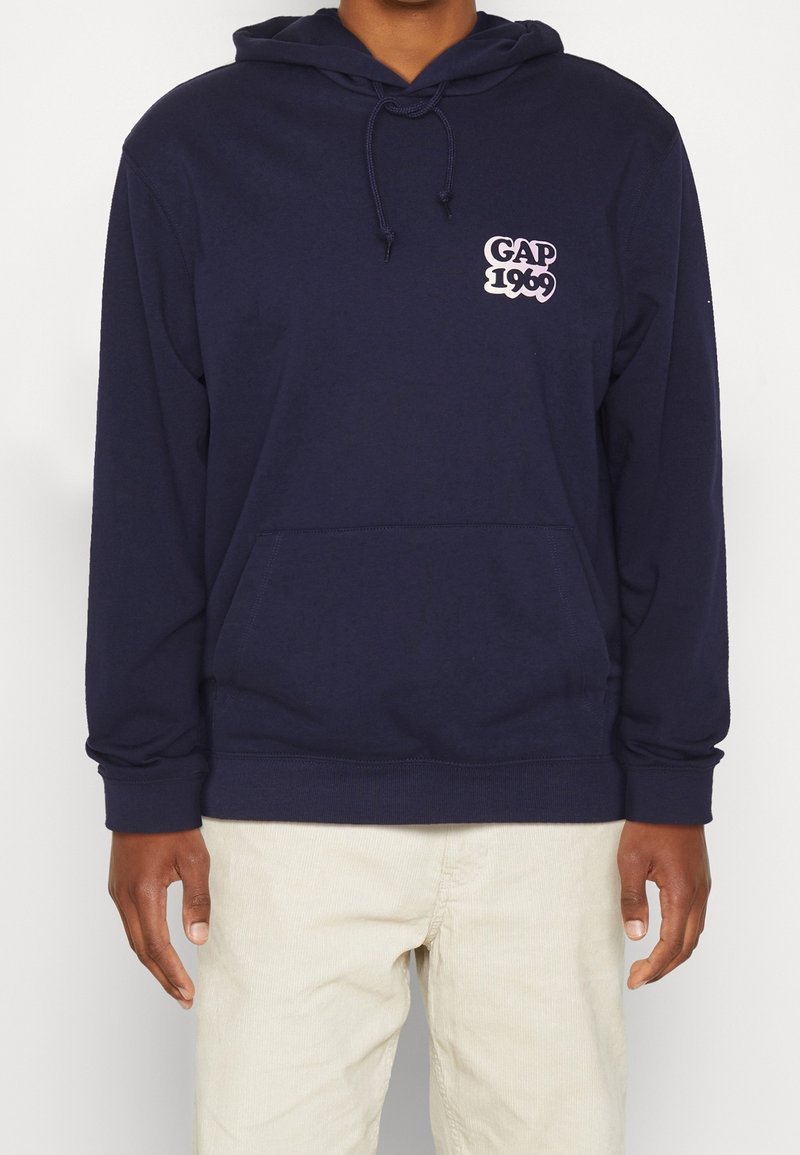 Personne portant un sweat à capuche bleu marine avec poche avant et logo "GAP 1969" sur la poitrine, associé à un pantalon beige clair.