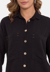 Zwarte button-up shirt van textuurstof, met twee borstzakken, houten knopen en contrasterende oranje stikselaccenten.