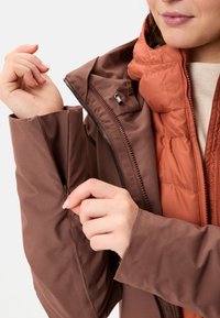 Bruine geïsoleerde jas met een ritsmanchet en een oranje binnenlaag. De buitenlaag is glad met een matte afwerking en biedt warmte.