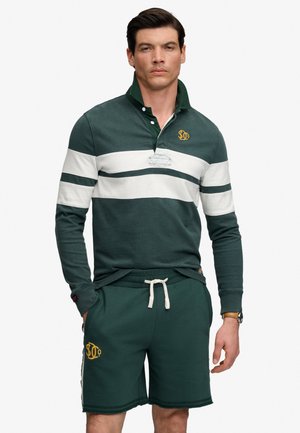 Mężczyzna ubrany w ciemnozieloną i białą pasiastą koszulkę do rugby z długim rękawem oraz pasujące zielone spodenki z żółtymi logo i zegarkiem na nadgarstku, stojący z rękami w kieszeniach.