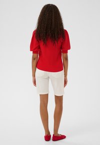 Haut rouge en maille à manches courtes associé à un short crème. Le haut présente des manches bouffantes et une coupe ajustée, tandis que le short arrive au niveau des genoux.