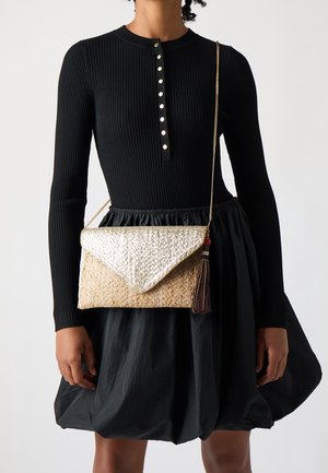 Kobieta ubrana w czarną, prążkowaną bluzkę z długim rękawem oraz spódnicę balonową, niosąca plecioną, złoto-beżową torebkę na ramię z frędzlami.