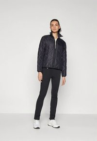 Žena v černé prošívané bundě, černých slim kalhotách a bílých teniskách, stojící na jednobarevném světlém pozadí.