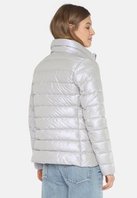 Silberne Pufferjacke mit horizontaler Steppung, hohem Kragen und glatter, glänzender Oberfläche. Kombiniert mit hellblauen Jeans.