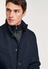 Merino villakangastakki tummansinisessä herringbone-kuosissa, jossa on kaksi nappia, korkea kaulus ja musta pehmustettu vuori. Varustettu vetoketjullisella sisävuorilla ja sivutaskuilla.