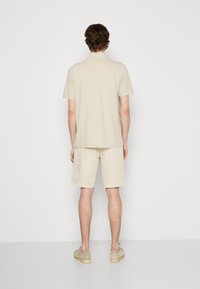 Polo-shirt beige à manches courtes et short assorti, texture douce, coupe décontractée, avec un design simple. Porté avec des chaussures de couleur claire.