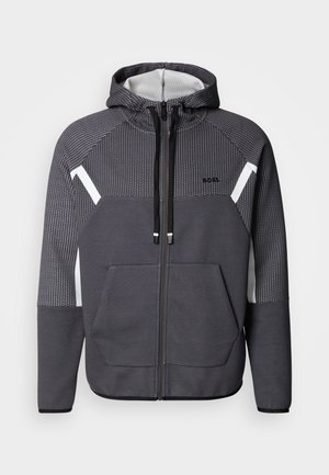 Corta-vento cinza com um padrão texturizado, detalhes brancos, dois bolsos frontais e capuz com cordão. Apresenta um design desportivo e moderno.
