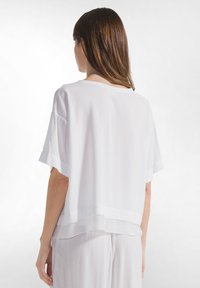Vrouw van achteren gezien, gekleed in een losvallende, witte top met korte mouwen en bijpassende witte broek tegen een effen witte achtergrond.