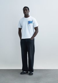 Mladý muž stojí s rukami v vreckách, oblečený v bielom tričku s potlačou, čiernych voľných džínsach a čiernych športových topánkach na šedej podlahe.