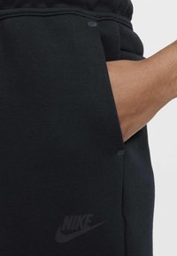 Pantalon de survêtement noir Nike doté d'une poche latérale, d'un tissu doux et texturé, avec un logo discret sur la partie inférieure de la jambe. Coutures apparentes.