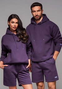 Les hoodies en coton violet présentent des cordons de serrage, des poignets côtelés et des poches kangourou, associés à des shorts assortis. Le tissu est lisse et décontracté.
