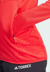 Maglia a maniche lunghe rossa con finitura testurizzata, dotata di tasca con zip sul lato, abbinata a pantaloni neri con il logo "TERREX".